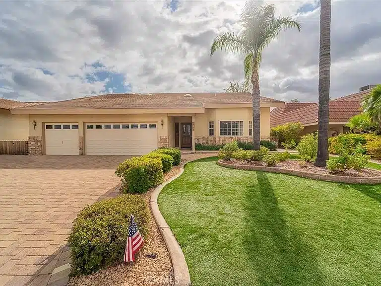 A single-story house with a three-car garage. The landscaped front yard features a lush green lawn, bushes, a curved sidewalk, and an American flag. The house has a beige exterior with large windows and palm trees in the yard under a partly cloudy sky.