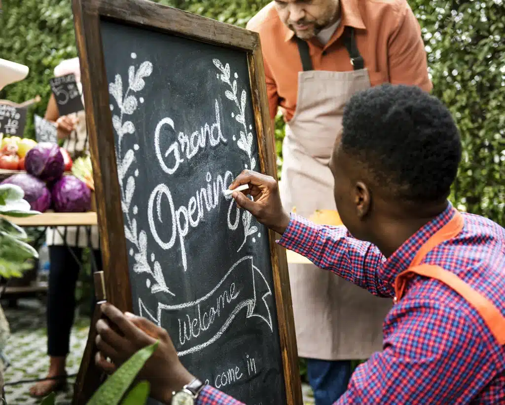 A person wearing a red checked shirt and orange apron writes "Grand Opening" on a chalkboard sign with white chalk. Another person in an apron stands behind, near a market stall with various vegetables like cabbages. Green foliage is visible around them.