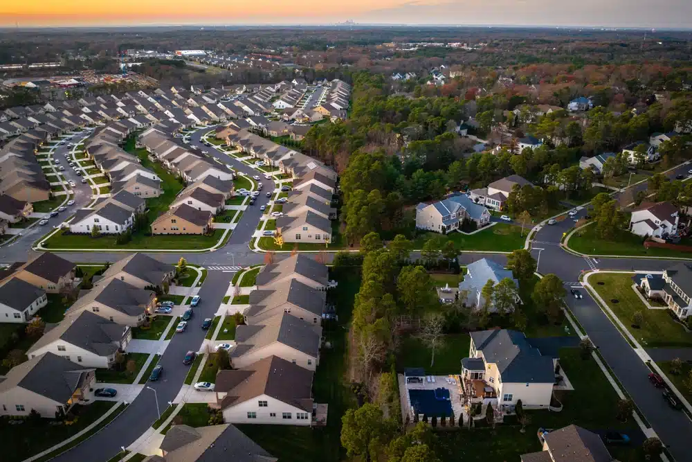 An aerial view at sunset of a suburban neighborhood with rows of houses featuring similar designs, landscaped lawns, and tree-lined streets. The image captures a mix of tightly clustered homes and larger properties with more expansive greenery and varied architecture.