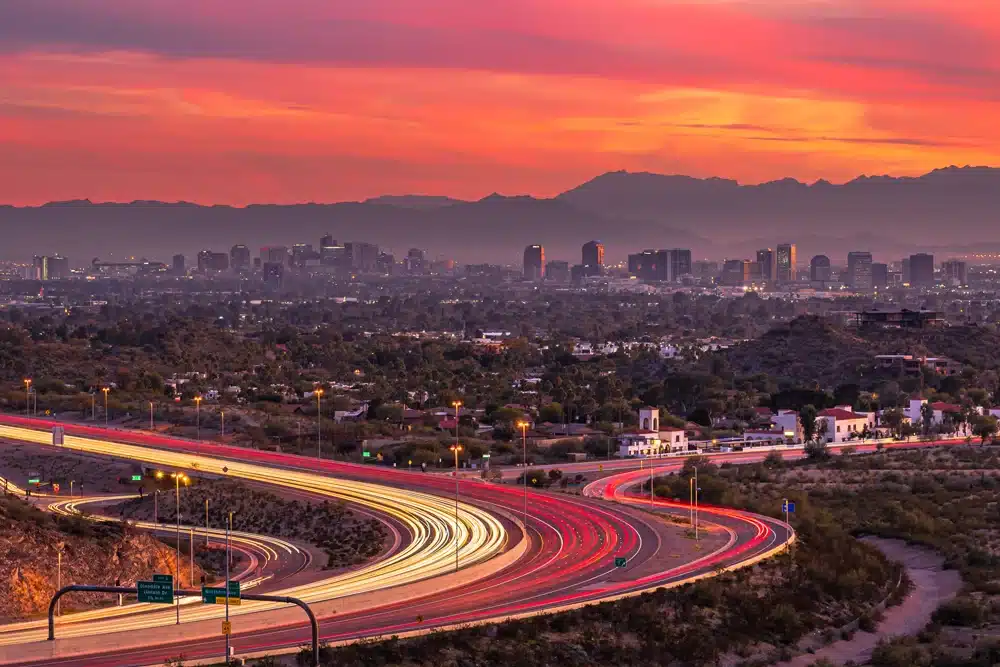 A vibrant sunset over a sprawling cityscape with distant mountains in the background. Bright, winding highways with streaks of car lights dominate the foreground, leading towards the city with tall buildings illuminated by the fading sunlight.