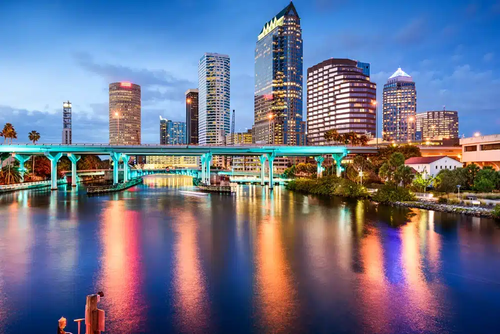 A vibrant cityscape with illuminated skyscrapers reflecting on a calm river at dusk. A colorfully lit bridge spans the river, with buildings of varying heights and designs creating a lively, urban atmosphere against a clear, twilight sky.