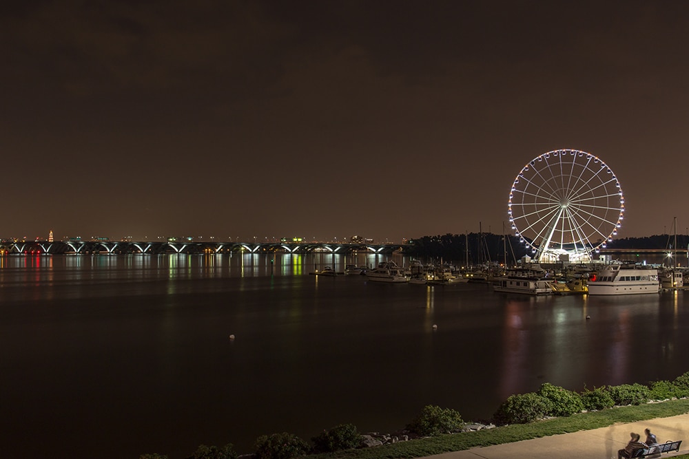 A nighttime view of a waterfront with a brightly lit Ferris wheel on the right. The Ferris wheel reflects on the calm water. Boats are docked nearby, and a bridge with lights is visible in the background. The sky is dark, and the area exudes a peaceful ambiance.