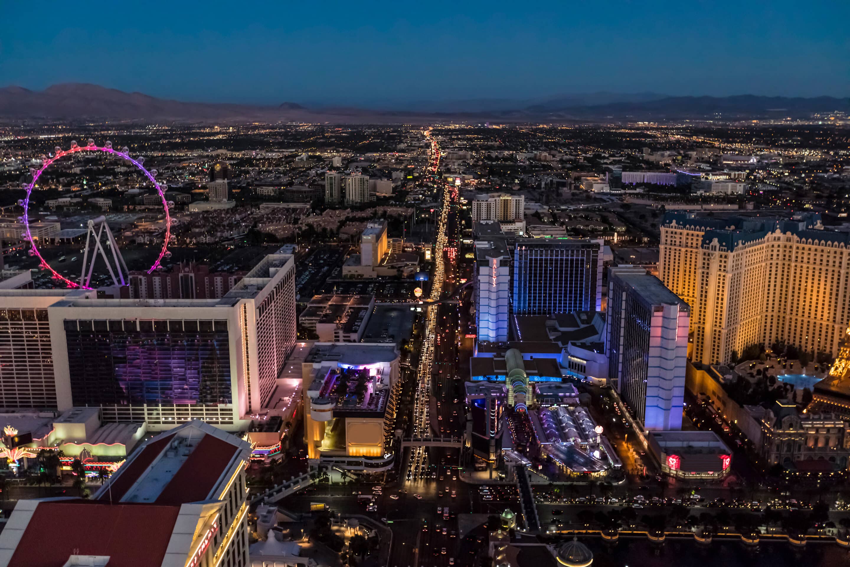 Aerial view of the Las Vegas Strip at dusk, showcasing brightly lit hotels and casinos. The High Roller Ferris Wheel is visible on the left, creating a striking contrast against the city's skyline. Mountains are faintly visible in the background, setting an exciting prelude to IDIQ at ASW 2025.