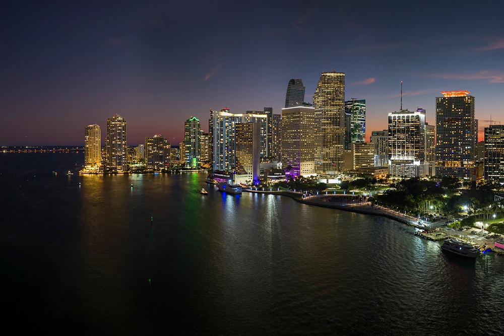 Aerial view of a brightly lit city skyline at night, featuring tall skyscrapers reflecting on the water. The dark sky is partially illuminated by fading daylight, creating a serene, urban waterfront scene.