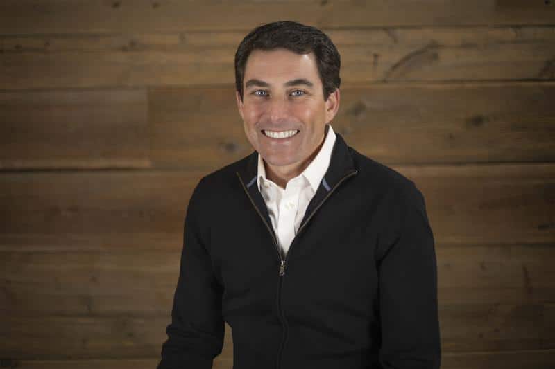 Bryan Sullivan, a man with short dark hair smiles at the camera. He is wearing a black zip-up sweater over a white collared shirt. The background is a wooden wall with horizontal planks.